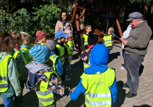 Grupa dzieci w odblaskowych kamizelkach stoi przed budynkiem Domu Pomocy Społecznej „Chemik”. W tle widać samochód i osobę dorosłą. Dzieci uczestniczą w akcji Klubu Małego Wolontariusza związanej z sadzeniem cebulek kwiatów dla pensjonariuszy domu opieki.