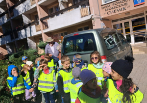 Grupa dzieci w odblaskowych kamizelkach słucha mężczyzny, który trzyma kartkę i czyta ułożone przez pensjonariuszy Domu Opieki podziękowania dla przedszkolaków. Obok stoi pedagog specjalny. Zajęcia odbywają się na zewnątrz, w ogrodzie przy Domu Pomocy Społecznej „Chemik”.