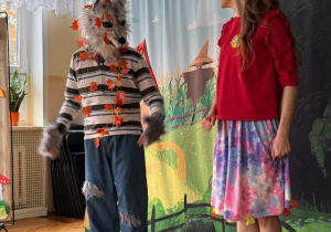 Aktor w masce wilka i aktorka w kolorowym stroju występują w przedszkolnym przedstawieniu na tle bajkowej scenografii z domkiem i dużymi kwiatami.