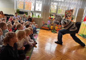 Dzieci w przedszkolu oglądają przedstawienie teatralne, w którym aktor w masce wilka występuje przed publicznością.