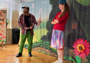 Dwójka aktorów występuje w przedszkolnym przedstawieniu na tle kolorowej scenografii z domkiem i dużym kwiatem