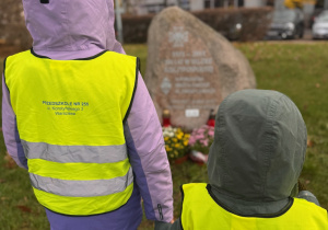 Dwójka dzieci w żółtych kamizelkach odblaskowych z napisem 'PRZEDSZKOLE NR 255, stoi przed kamiennym pomnikiem pamięci. Dzieci trzymają się za ręce i patrzą na pomnik, wokół którego ustawiono kwiaty i znicze. W tle widoczne są budynki mieszkalne oraz drzewa z jesiennymi liśćmi.