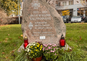 Kamienny pomnik upamiętniający 100-lecie służby 16. Warszawskiej Brygady Pancernej im. Zawiszy Czarnego. Pomnik stoi na trawie, w tle widoczne są budynki mieszkalne. Na kamieniu wygrawerowany jest tekst rocznicowy oraz emblemat wojskowy. Przed pomnikiem ustawiono donice z kwitnącymi chryzantemami i dwa czerwone znicze – symbole pamięci i szacunku.”