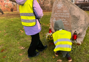 Dwoje dzieci w żółtych kamizelkach odblaskowych składa kwiaty i zapalone znicze pod kamiennym pomnikiem na trawie. Na pomniku widoczny jest napis upamiętniający 100-lecie 16 Warszawskiej drużyny harcerskiej. W tle znajdują się drzewa i fragment budynku.”