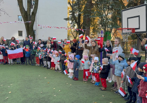 Dzieci z przedszkola stoją wraz z rodzicami i opiekunami na przedszkolnym placu podczas uroczystości patriotycznej. Cała grupa trzyma w rękach biało-czerwone flagi, a niektóre dzieci prezentują dużą flagę Polski. Uczestnicy ustawieni są w długim szeregu, uśmiechnięci i zaangażowani we wspólne śpiewanie hymnu w ramach obchodów ‘Szkoła do hymnu’. W tle widać dekoracje z biało-czerwonych girland oraz otoczenie placu zabaw.