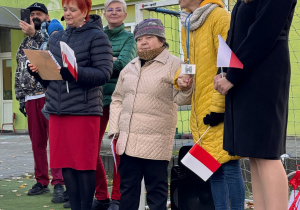 Uczestnicy wydarzenia- pracownicy przedszkola oraz przedstawiciele i pensjonariusze Domu Pomocy Społecznej ‘Chemik’- biorą udział we wspólnym spotkaniu patriotycznym. Jedna z osób (Pani dyrektor przedszkola) trzyma w rękach kartkę z podziękowaniami od pensjonariuszy, a inni trzymają biało-czerwone flagi. Wszyscy stoją w szeregu, uśmiechają się i uczestniczą w uroczystej części wydarzenia.”
