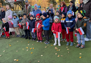 Grupa przedszkolaków stoi razem z opiekunami na przedszkolnym placu podczas uroczystości patriotycznej. Dzieci trzymają w rękach biało-czerwone flagi i ustawione są w szeregu, ubrane w ciepłe kurtki i czapki. W tle widać dorosłych uczestników wydarzenia oraz elementy placu zabaw. Cała grupa bierze udział we wspólnym śpiewaniu hymnu w ramach akcji ‘Szkoła do hymnu’