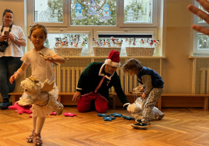 Dwoje dzieci wykonujących zadanie dla mikołaja, w tle zdjęcia widać mężczyznę w ubraniu pomocnika Świętego Mikołaja.