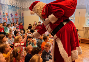 Święty Mikołaj wita się z dziećmi, przybijajcąc piątki z dziećmi siedzącymi na dywanie.