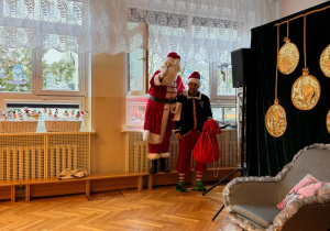 po prawej stronie zdjęcia Mężczyzna w stroju Świętego Mikołaja wchodzi do przedszkola przez okno, Elf pomaga mu w tym podając mu rękę.