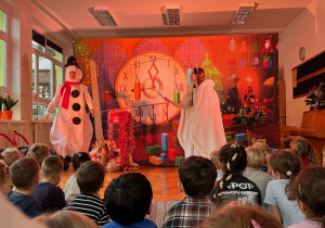 Aktor przebrany za bałwanka oraz aktorka w stroju Królowej Śniegu występują na scenie, a na pierwszym planie widoczne są głowy dzieci oglądających przedstawienie.