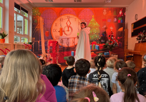 Dzieci z przedszkola oglądają przedstawienie teatralne Elfy i fabryka prezentów w świątecznej scenerii. Na scenie przed dziećmi stoi zła królowa śniegu w białej pelerynie.