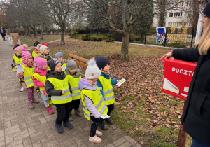 Dzieci z grupy II w czapkach zimowych kurtkach i kamiezelkach odblaskowych z logiemprzedszkola ustawiają się przed skrzynką pocztową aby wrzycić wykonaną kartkę do skrzynki pocztowej.