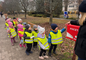 grupa stoi za chłopcem w niebieskiej kurtce i granatowej czapce, który wrzuca do skrzynki pocztowej własnoręcnie wykonaną kartkę świąteczną.