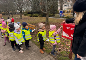Dziewczynka w jasno różowej czapce oraz różowej kurtce wrzuca do skrzynki kartkę świąteczną, reszta grupy czeka na swoją kolej.