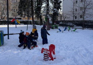 dzieci bawią się na zaśnieżonym placu zabaw, bujają się na bujanym koniku i ciągną wzajemnie na saneczkach.