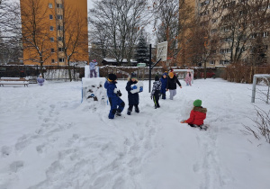 chłopcy z grupy czwartej na zaśnieżonym boisku przedszolnym bawią się w śniegu i wspólnie próbują ulepić bałwana.