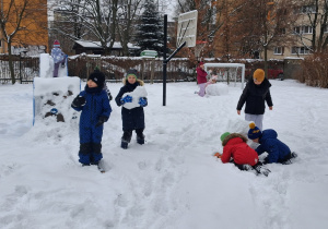 Na zaśnieżonym placu zabaw widać na pierwszym planie chłopców starających się złożyć bałwana z utoczonych wcześniej kul, natomiast na drugim planie dziewczynki toczą wspólnie dużą kulę ze śniegu.
