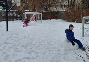 Na zaśnieżonym boisku przedszkolnym na pierwszym planie jeden z chłopców z grupy czwartej siedzi na śnieżnej kuli zaś na drugim planie, dwie dziewczynki lepią kulę śniegu.