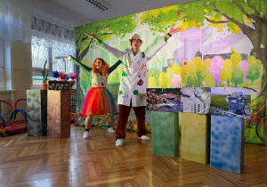 Dwójka aktorów podczas kolorowego spektaklu ekologicznego w sali przedszkolnej. Kobieta w tęczowej spódnicy i mężczyzna w białym fartuchu stoją na tle dekoracji przedstawiającej park i unoszą ręce do góry.