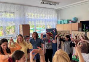 Grupa kobiet oraz jedna dziewczynka w stroju tanecznym tworzy koło i tańczy, trzymając się za ręce, w jasnej, udekorowanej klasie szkolnej. W tle widoczne są dekoracje o tematyce wiosennej oraz okna z firankami. Atmosfera na zdjęciu jest radosna i zaangażowana.